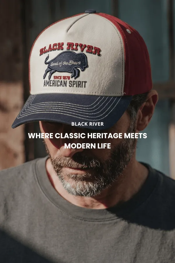 Een man zit op een houten kist in een studio en draagt een klassieke bruine cowboyhoed van vilt met brede rand, een donker denim overhemd en een spijkerbroek.