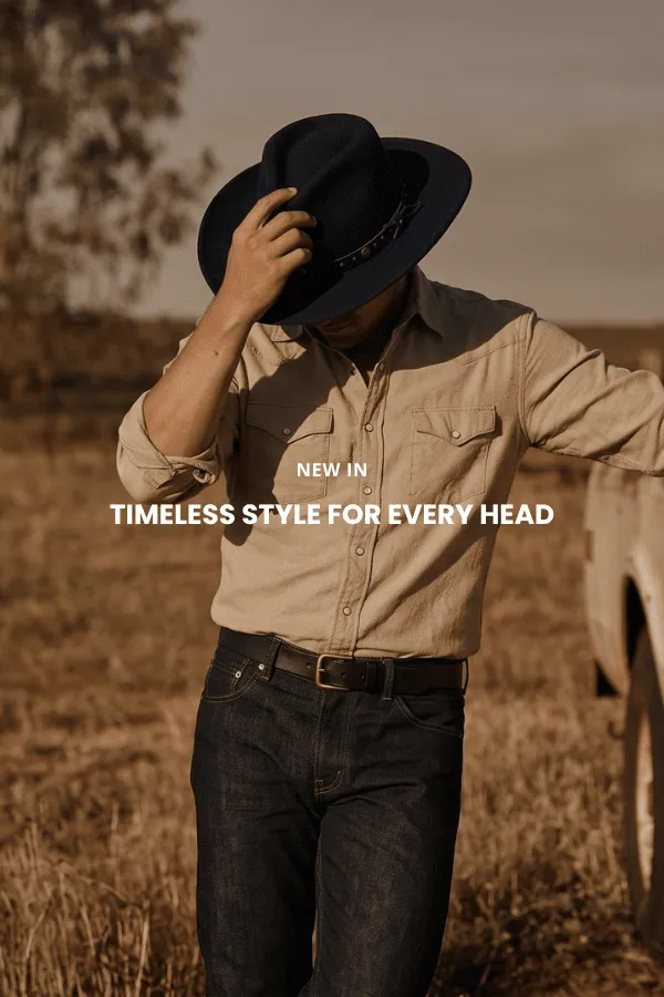 Een man zit op een houten kist in een studio en draagt een klassieke bruine cowboyhoed van vilt met brede rand, een donker denim overhemd en een spijkerbroek.