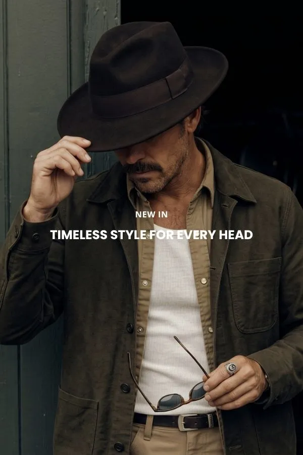 Een man zit op een houten kist in een studio en draagt een klassieke bruine cowboyhoed van vilt met brede rand, een donker denim overhemd en een spijkerbroek.